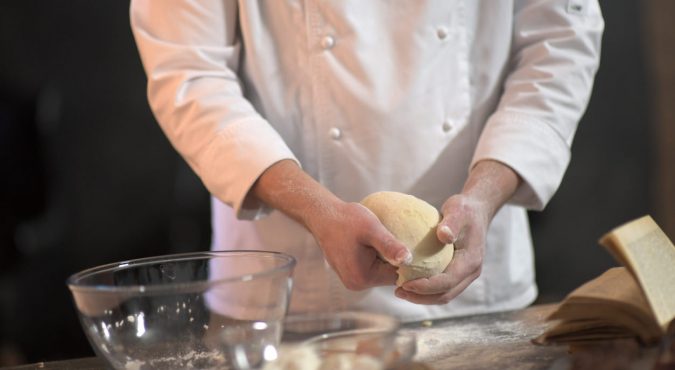 Cook hands kneading dough, sprinkling piece of dough with white wheat flour. Low key shot, close up on hands, some ingredients around on table. An experienced chef in a professional kitchen prepares the dough with flour to make the bio Italian pasta. the
